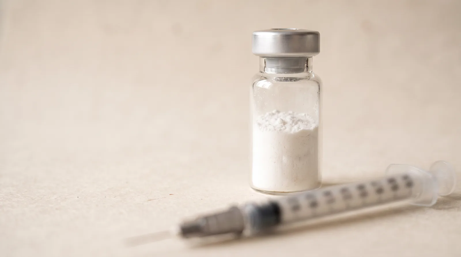 Illustration: a small unlabelled glass vial of powder in focus with an insulin syringe blurred in the foreground.